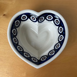 May include: A heart-shaped ceramic bowl with a white interior and a blue rim. The rim is decorated with a repeating pattern of blue circles with red and white centers. The bowl is likely used for serving or display.