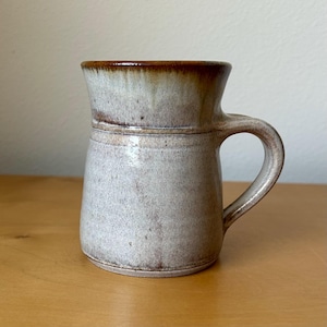 May include: A ceramic mug with a speckled light gray body and a smooth, glossy brown glaze around the rim. The mug features a wide, flared top section, a slightly tapered cylindrical base, and a sturdy, curved handle attached to the side.