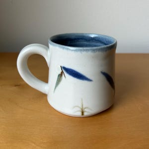 May include: A white ceramic mug with a blue rim and handle. The mug is decorated with blue and green leaf-like designs. The mug is sitting on a wooden surface.