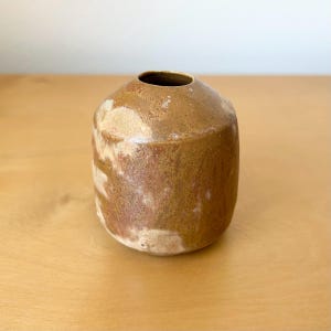 May include: A small, cylindrical ceramic vase with a rounded top and a narrow circular opening. The exterior features a mottled, earthy glaze with blended tones of tan, brown, and cream, creating a textured, variegated appearance across the surface.