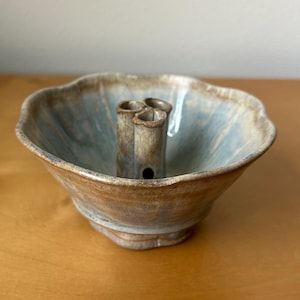 May include: A handmade ceramic bowl with a light blue interior and a brown exterior. The bowl has a unique design with three cylindrical holders in the center. The rim of the bowl has a wavy, organic shape. This decorative piece is ideal for home decor.