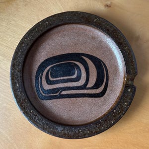 May include: A round, brown ceramic ashtray with a dark brown rim and a central design in black. The design is an abstract, nested shape. The ashtray has a small notch on the side.