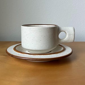 May include: A speckled white ceramic coffee cup and saucer set. The cup has a unique rectangular handle with a circular opening. The rim and the edge of the saucer are trimmed with a brown band. The set is on a wooden surface.