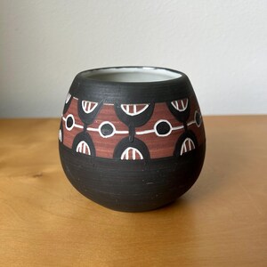 May include: A round ceramic vessel with a matte black finish featuring a wide horizontal band of terracotta red. The band is decorated with a repeating pattern of black and white semi-circular arches and small white circles encircling the exterior.