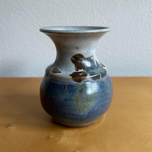 May include: A small, handcrafted ceramic vase with a rounded base and flared neck. The vase features a color palette of blue, brown, and white, with a glossy finish. The vase is suitable for displaying small flowers or as a decorative accent.