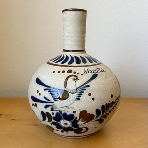 May include: A ceramic vase with a round body and a long neck. The vase is beige with a brown rim and features a hand-painted bird and floral design in blue and brown. The word "Mazatlan" is written on the vase.