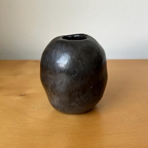 May include: A small, rounded vase with a smooth, dark charcoal-black finish. The vessel features an organic, slightly asymmetrical shape with a narrow circular opening at the top and a subtle, mottled texture across its polished, dark surface.