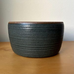 May include: A handcrafted, dark blue ceramic bowl with a textured surface and a brown rim. The bowl has a wide, rounded shape and is suitable for serving or display. The exterior features horizontal, ridged detailing.