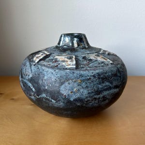 May include: A squat, round ceramic vase with a dark, mottled black and blue glaze. The surface features a textured, rugged finish with lighter, off-white geometric patterns radiating from the narrow, circular opening at the top of the vessel.