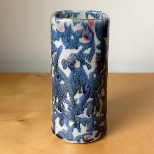 May include: A cylindrical ceramic vase with a square top. The vase features a blue, white, and maroon pattern. The glaze has a dripped effect, with hints of gold. The vase is on a wooden surface.