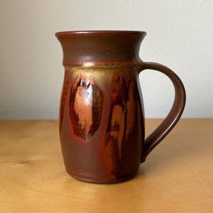 May include: A ceramic mug featuring a flared rim and a curved handle. The exterior displays a glaze with blended tones of deep brown, burnt orange, and golden yellow, creating a vertical drip pattern that transitions into a solid dark brown top.