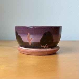 May include: A small, handcrafted ceramic planter with a desert landscape design. The planter features a deep purple exterior with a brown base and a terracotta-colored saucer. The design includes a cactus and mountain silhouettes.