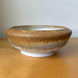 May include: A round ceramic bowl featuring a wide, thick rim with a glossy brown glaze that transitions into a lighter, mottled tan and white finish toward the base. The interior of the bowl is smooth and white, while the exterior base is white.