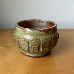 May include: Handmade ceramic bowl with a textured, carved design. The bowl features a green exterior with brown and white accents around the rim. The interior is a light brown color. This unique pottery piece is perfect for serving or display.