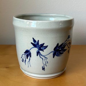 May include: A light gray ceramic planter with a blue floral design. The planter has a cylindrical shape with a slightly flared rim and a base. The blue floral design features leaves and small clusters of berries.