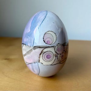 May include: A decorative egg-shaped art piece with a glossy finish. The egg features a design with pastel colors, including light blue, pink, and gray, with circular patterns and black outlines. The overall aesthetic is artistic and unique.