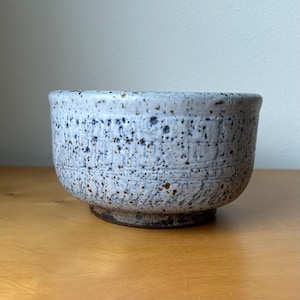 May include: A ceramic bowl with a speckled white and light gray glaze featuring a textured, impressed pattern around the exterior. The base of the bowl is a contrasting dark brown, unglazed clay, providing a stable foundation for the vessel.