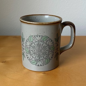 May include: A ceramic mug with a light gray speckled finish and a brown rim and handle. The mug features a central green and black floral design. The mug is suitable for hot or cold beverages.