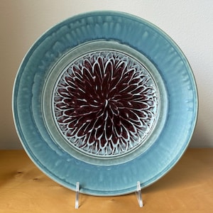 May include: A round, ceramic plate with a teal blue rim and a central design. The center features a maroon floral pattern with white accents. The plate is displayed on a clear stand, showcasing its decorative design.