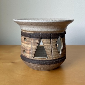 May include: A handcrafted ceramic candle holder with a wide, speckled rim and geometric cutouts. The pottery features a mix of brown, tan, and cream tones, with dark brown bands and triangular and diamond-shaped openings. The candle holder is designed to hold a candle.