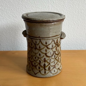 May include: A ceramic jar with a lid, featuring a light brown and tan color scheme. The jar has a cylindrical shape with a textured, patterned design in brown. Two small handles are on the sides. The lid sits on top, completing the design.