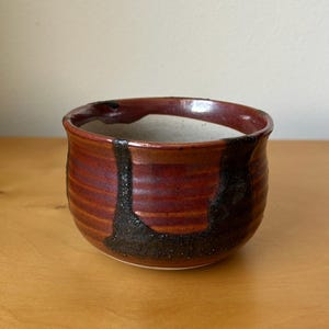 May include: A handmade ceramic bowl with a reddish-brown exterior and a white interior. The bowl features a dark, textured, vertical design element. The bowl is round with a slightly flared rim and sits on a light-colored surface.