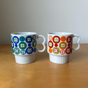 May include: Two white ceramic mugs with a stacked design. One mug features blue, teal, and green circles with flower-like patterns. The other mug has orange, red, and green circles with similar flower patterns. Both mugs have heart-shaped handles.