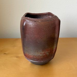 May include: A hand-thrown ceramic vase with a deep reddish-brown and charcoal gradient finish. The vessel features a square-shaped opening with rounded corners and subtle horizontal ridged detailing along the top rim, showcasing a textured surface.