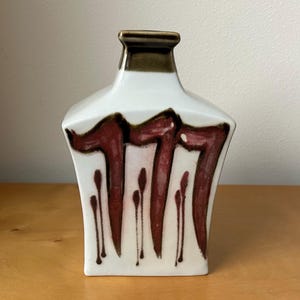 May include: A white ceramic vase with a rectangular shape and a dark brown rim. The vase features a bold, abstract design in shades of burgundy and brown, with vertical drips. The vase is displayed on a wooden surface.