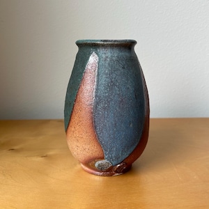 May include: A ceramic vase with a tapered cylindrical shape, featuring a textured surface with a blend of deep teal, slate blue, and earthy terracotta brown glazes. The finish shows subtle variations in color and a speckled, matte appearance.