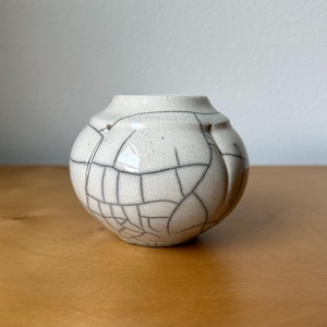 May include: A small, round ceramic vase with a cream-colored glaze and a prominent black crackle pattern across its surface. The vessel features a wide, rounded body that tapers toward a narrow, open top, showcasing a distinct network of fine lines.