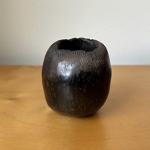 May include: A small, rounded, dark brown ceramic vessel with a smooth, polished finish on the lower body and a textured, grooved surface around the circular opening at the top. The object has a deep, dark coloration and a thick, uneven rim.