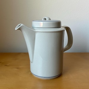 May include: A light gray ceramic teapot with a lid and handle. The teapot has a spout and a dark blue trim around the base and lid. The teapot is sitting on a wooden surface.