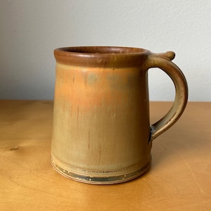 May include: A handmade ceramic mug with a unique handle. The mug features a gradient of colors, transitioning from a light tan to a warm orange and brown at the rim. The mug has a rustic, textured appearance, perfect for enjoying hot beverages.