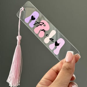 May include: A clear rectangular acrylic bookmark featuring the name Aria in a wavy, multi-toned pink and purple font. The design includes two small black heart accents and a light pink decorative tassel attached to the top corner with a cord.