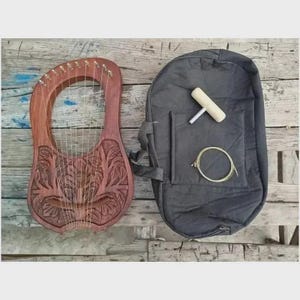 Może przedstawiać: Drewniana lira z rzeźbionymi wzorami kwiatowymi, obok szarej torby transportowej. Lira ma struny i czerwonobrązowe wykończenie. Etui zawiera narzędzie do strojenia i zapasową strunę. Tło to zwietrzała drewniana powierzchnia.