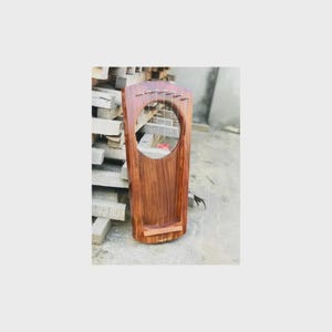 Può includere: Una lira in legno fatta a mano con sei corde. Lo strumento presenta una grande buca circolare e un corpo rettangolare. Il legno è di un ricco colore marrone scuro con venature visibili. La lira è progettata per suonare musica.