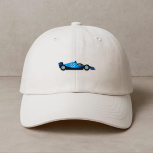 Może przedstawiać: Biała czapka z daszkiem z niebieskim wzorem samochodu wyścigowego i numerem 55. Czapka ma zakrzywiony daszek i regulowany pasek. Klasyczny dodatek dla entuzjastów sportów motorowych.