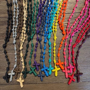 Op de afbeelding: Een verzameling kleurrijke rozenkranskettingen. Elke ketting heeft een geknoopt koord met kralen en een kruisvormige hanger. Kleuren zijn onder meer zwart, bruin, beige, groen, paars, blauw, geel, oranje, roze en rood. De kettingen worden op een houten oppervlak gepresenteerd.