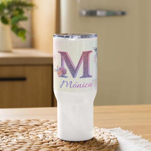 Pode incluir: Um copo de aço inoxidável branco com uma letra M grande, brilhante e com gradiente nas cores rosa, roxo e azul. Abaixo da letra, o nome Mónica está impresso em uma fonte cursiva roxa, acompanhado por gráficos de flores e corações.