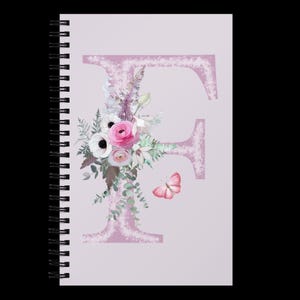 May include: A spiral-bound notebook with a light purple cover featuring a large, textured letter "F". A floral arrangement with pink roses, white anemones, and greenery adorns the letter. A pink butterfly is also present.