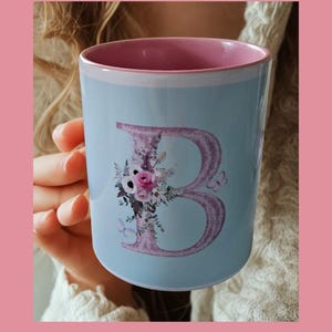 Pode incluir: Uma caneca de cerâmica azul-clara com interior rosa. O exterior apresenta uma letra B grande, texturizada e roxa, decorada com um arranjo floral de rosas cor-de-rosa, flores brancas e pequenas folhas verdes, acompanhada por duas pequenas borboletas.