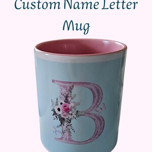 Pode incluir: Caneca de cerâmica azul clara com interior rosa e borda branca. A caneca apresenta um design floral da letra "B" com borboletas. O texto "Custom Name Letter Mug" é exibido em verde-azulado.