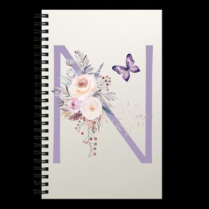 Puede incluir: Cuaderno de espiral con una cubierta crema. La letra "N" lavanda se muestra con un arreglo floral de acuarela y una mariposa en tonos rosas, morados y verdes. Perfecto para tomar notas.