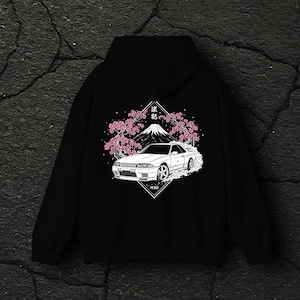 Op de afbeelding: Zwarte hoodie met een witte afbeelding van een auto, berg en kersenbloesem ontwerp. De afbeelding bevat Japanse tekst en het automodel R32. Het ontwerp is gecentreerd op de achterkant van de hoodie.