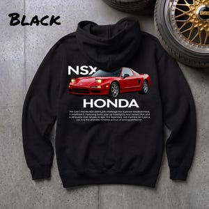 May include: A black hooded sweatshirt featuring a graphic of a red Honda NSX sports car with pop-up headlights. The text reads NSX HONDA followed by a paragraph describing the vehicle's history and impact on the supercar establishment.