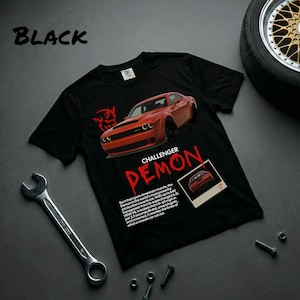 Può includere: T-shirt nero con grafica rossa Dodge Challenger Demon. La maglietta mostra la scritta "CHALLENGER DEMON" e informazioni sull'auto. Una chiave inglese, bulloni e un pneumatico sono visibili sullo sfondo.