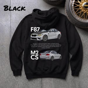 Puede incluir: Sudadera con capucha negra con ilustraciones de coches deportivos blancos y grises, con las etiquetas "F87" y "M2 CS". La sudadera también tiene la palabra "BLACK" en la parte superior y una descripción de las características del coche.