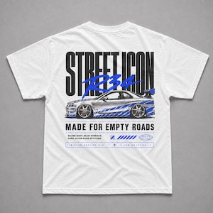 May include: White t-shirt featuring a graphic of a silver car with blue accents and the text "STREET ICON R34". Below the car, the text reads "MADE FOR EMPTY ROADS". Additional text includes "SILVER BODY. BLUE STREAKS. PURE AFTER-DARK ATTITUDE."