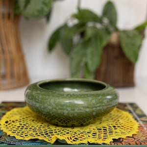 May include: A round, moss green ceramic bowl with a speckled glaze finish. The bowl sits atop a bright yellow circular crochet doily, which is placed on a hardcover book titled Jungalow by Justina Blakeney with a dark green spine and cover.